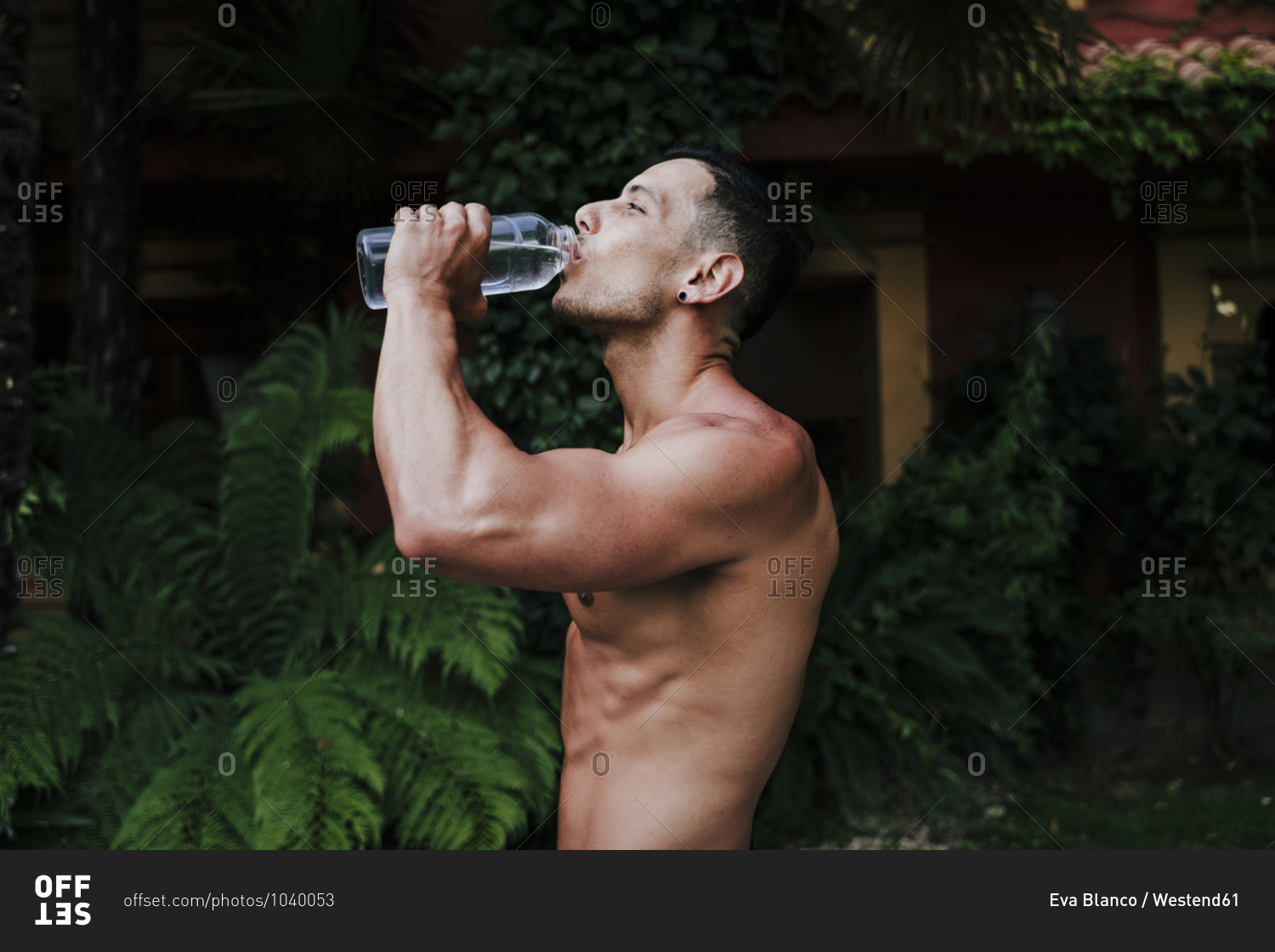 Shirtless male athlete drinking water while standing against plants in yard