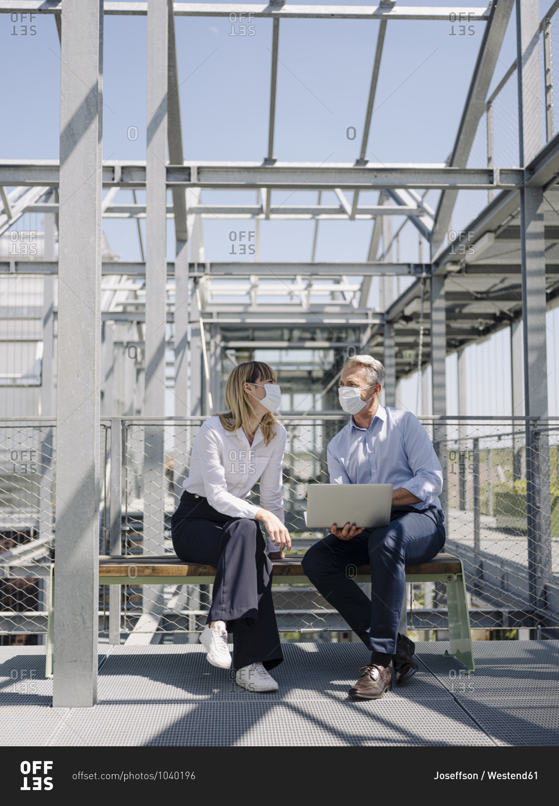 Business people wearing masks discussing over laptop while sitting on seat in greenhouse