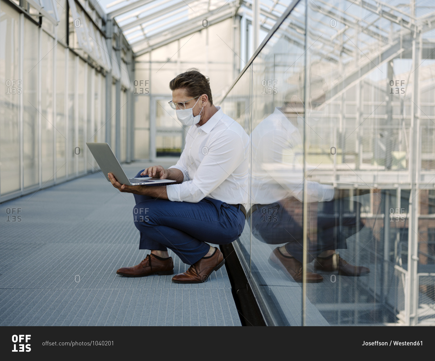 Businessman wearing mask using laptop while crouching on floor in greenhouse