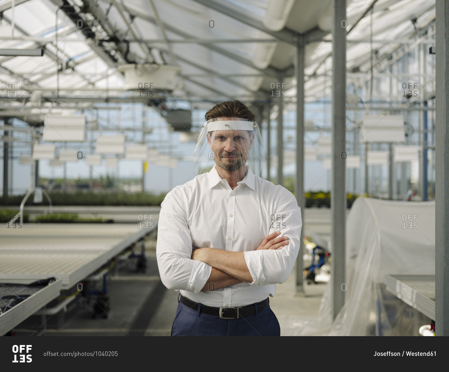 Businessman with arms crossed wearing face shield while standing in greenhouse