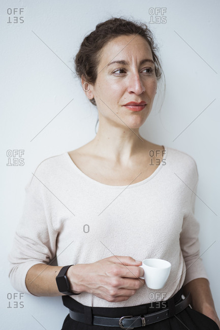 Thoughtful female entrepreneur holding coffee cup standing against white wall