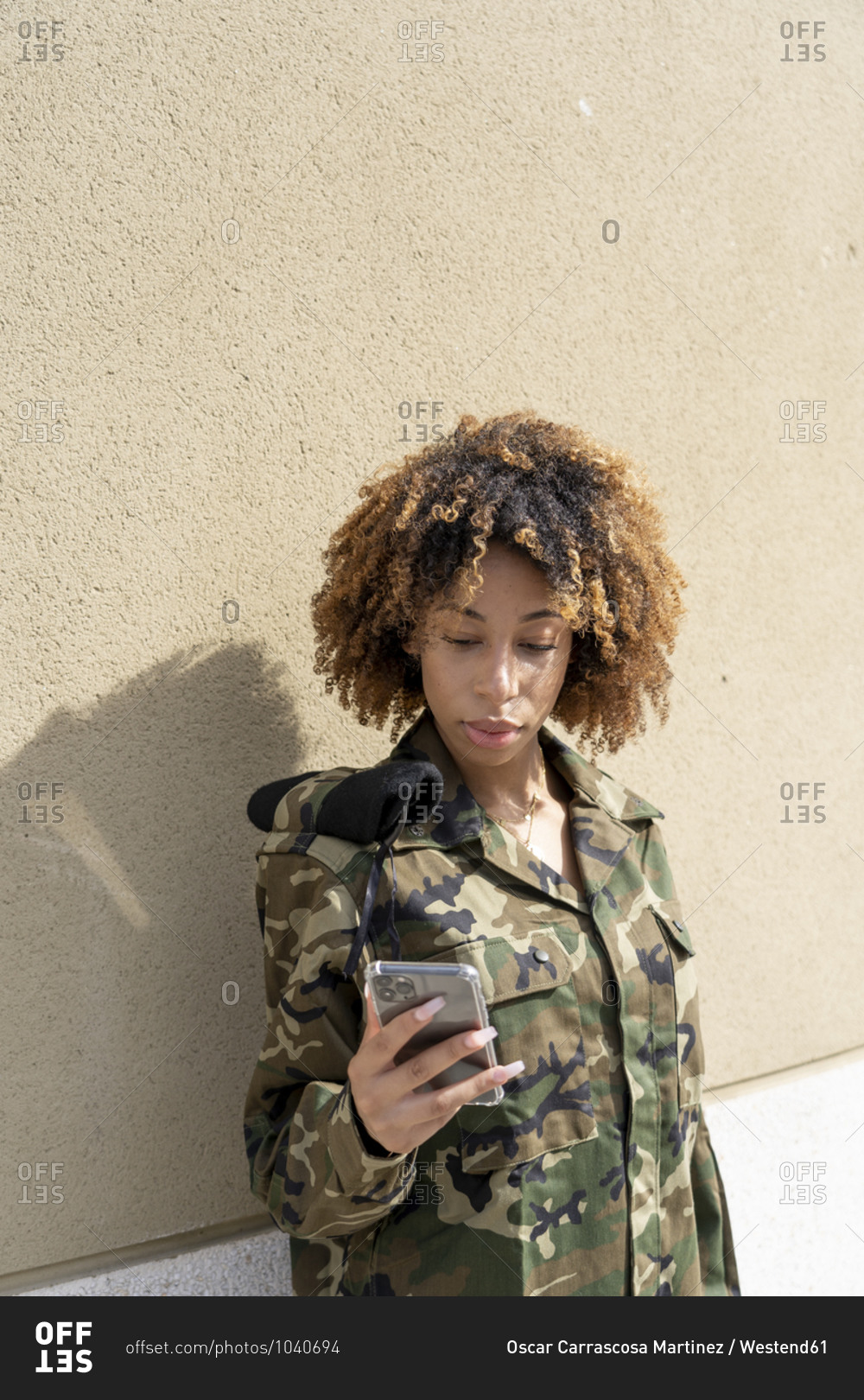 Female army soldier text messaging on smart phone while standing against wall