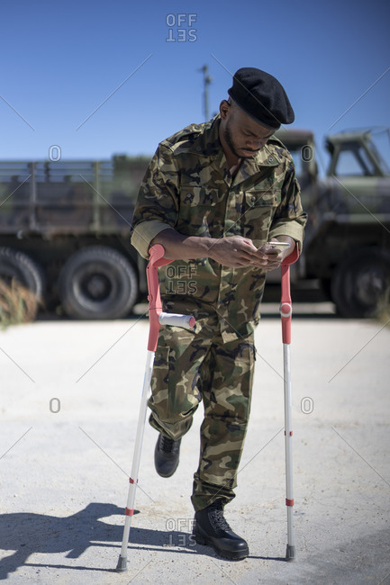 Injured army soldier with crutches text messaging on smart phone during sunny day