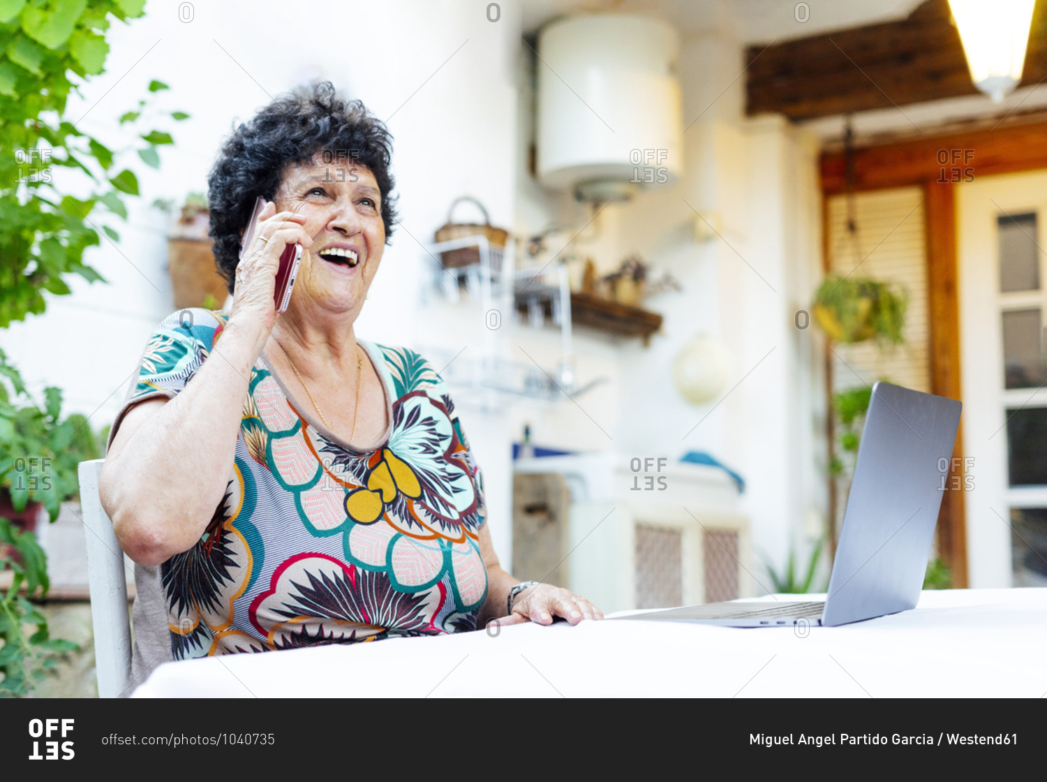 Cheerful senior woman talking over smart phone while sitting by laptop in yard