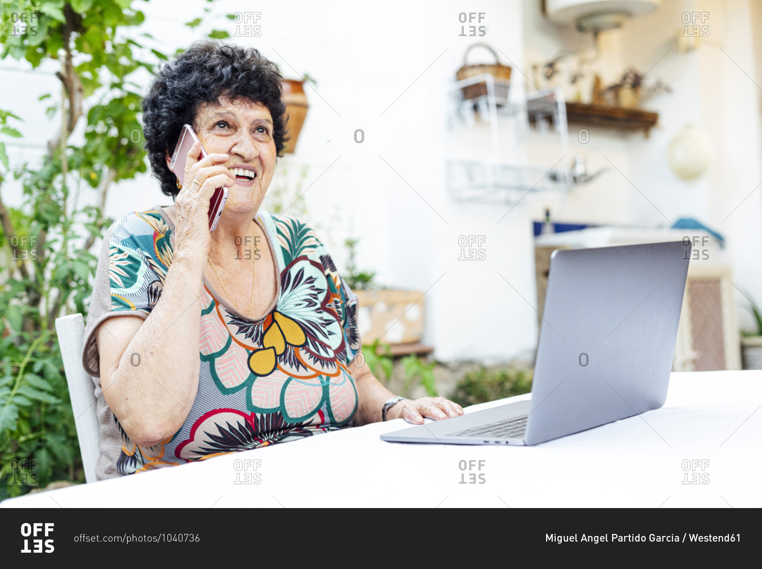 Happy senior woman with laptop on table talking over mobile phone in yard