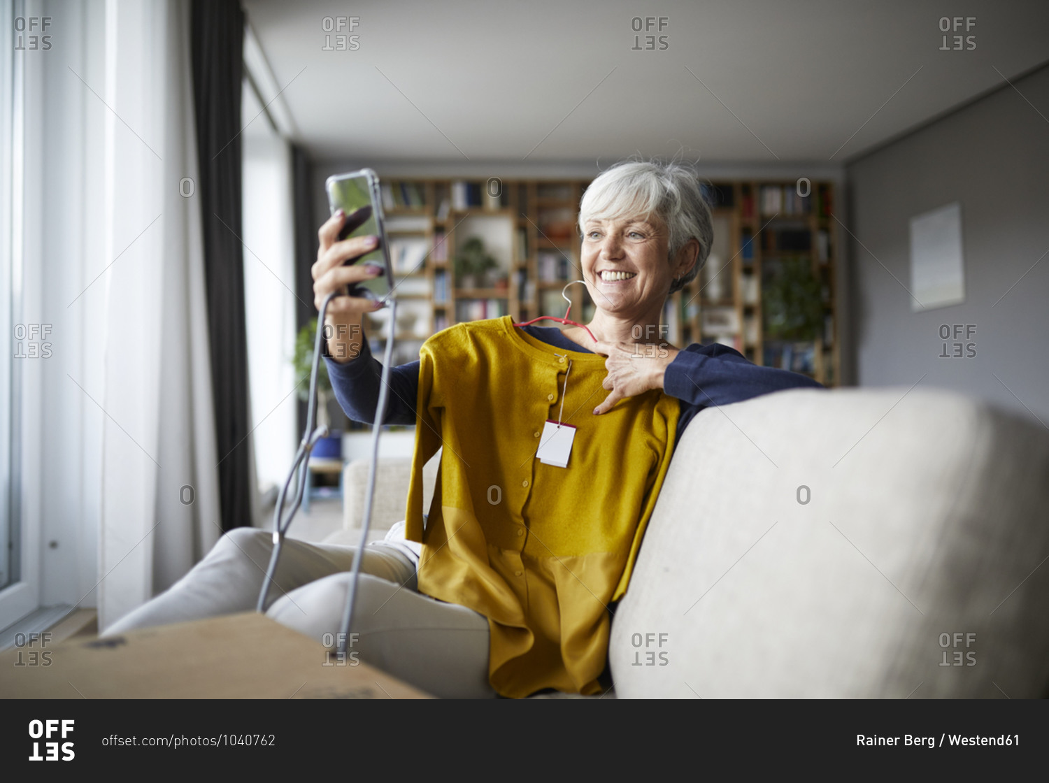Smiling senior woman taking selfie with new blouse on smart phone while sitting at home