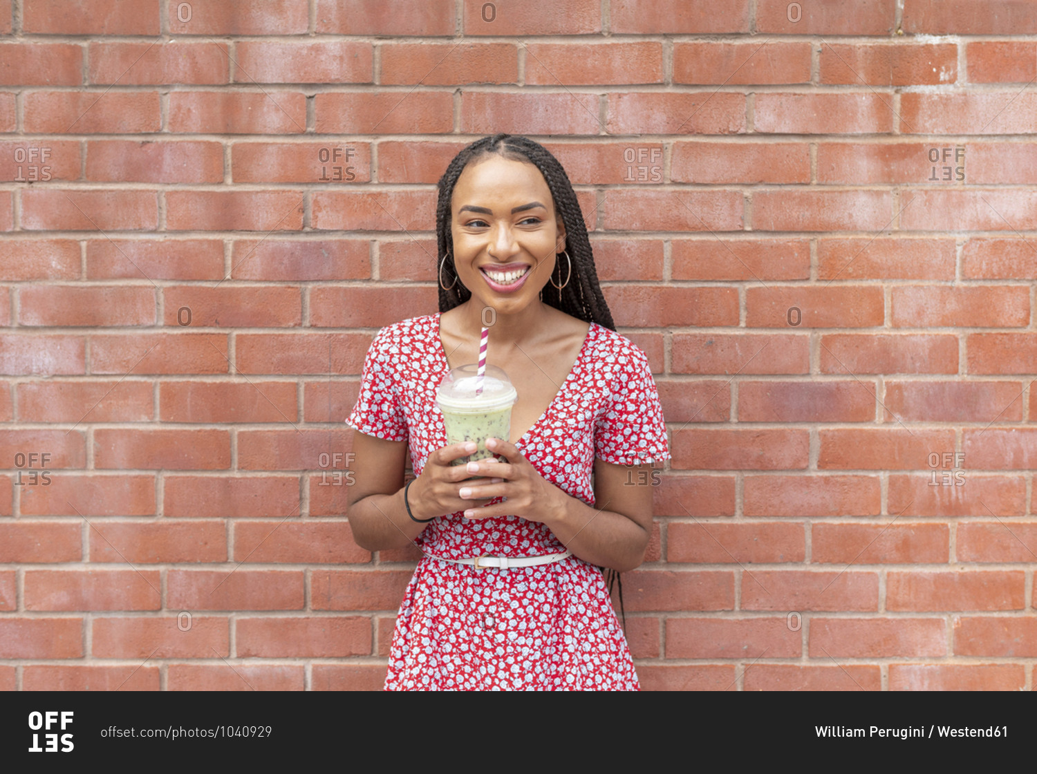 Young woman drinking smoothie while standing against brick wall