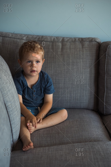 Little boy with bandage on forehead sitting on gray sofa