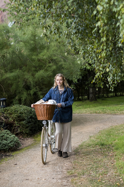 Young blond woman pushing bicycle with basket along footpath.