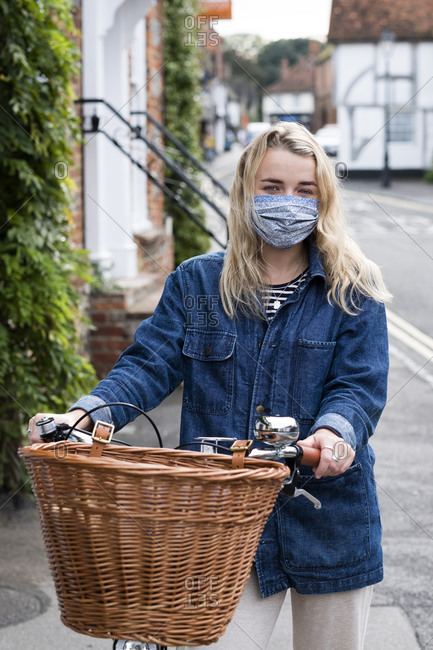 Young blond woman wearing face mask on bicycle with basket, looking at camera.