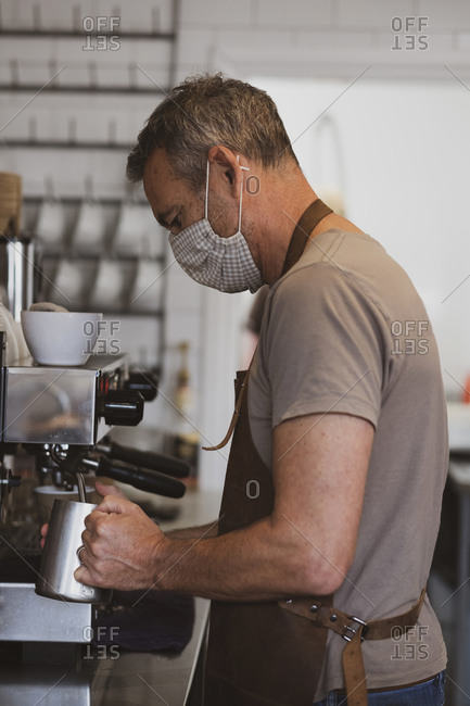 Male barista wearing brown apron and face mask working in a cafe, frothing milk.