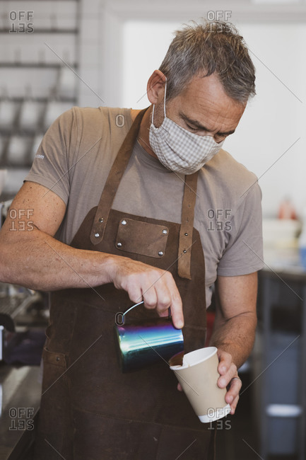 Male barista wearing brown apron and face mask working in a cafe, pouring coffee.