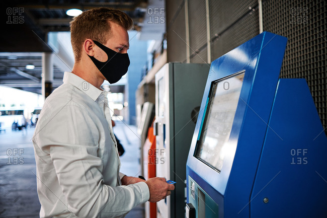 Side view of male wearing protective mask using modern self service terminal and touching display during coronavirus epidemic