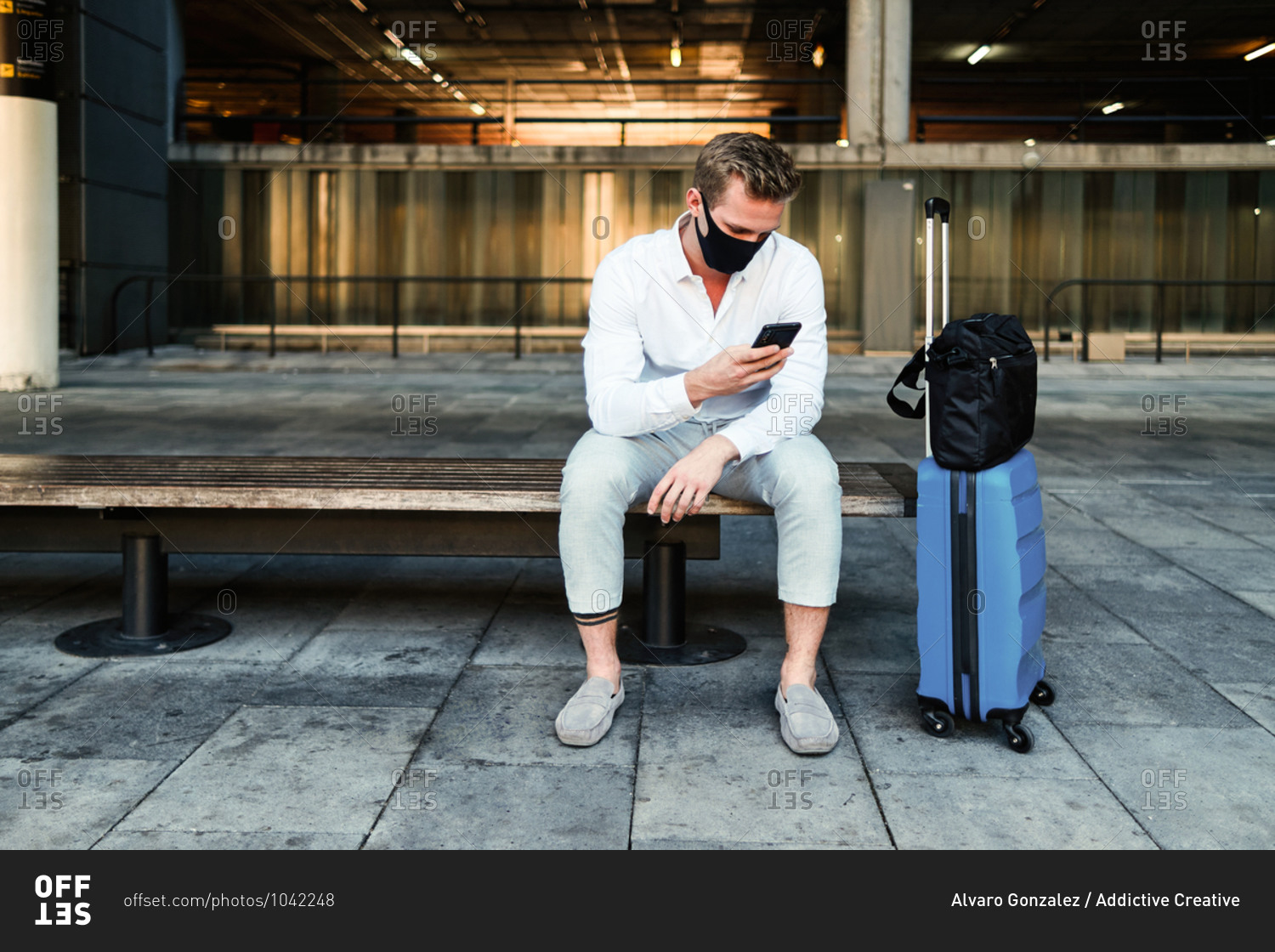 Calm male tourist in protective mask and with luggage sitting on bench in airport and using smartphone before departure during coronavirus pandemic
