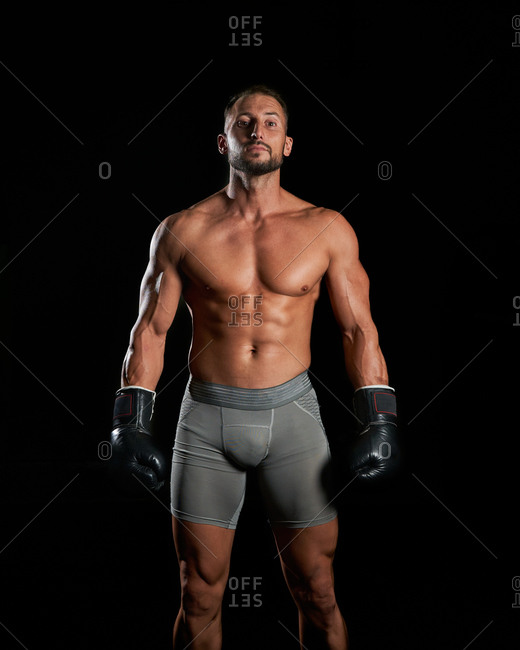 Brutal shirtless sportsman with muscular body with boxing gloves while preparing to fight standing looking at camera against black background