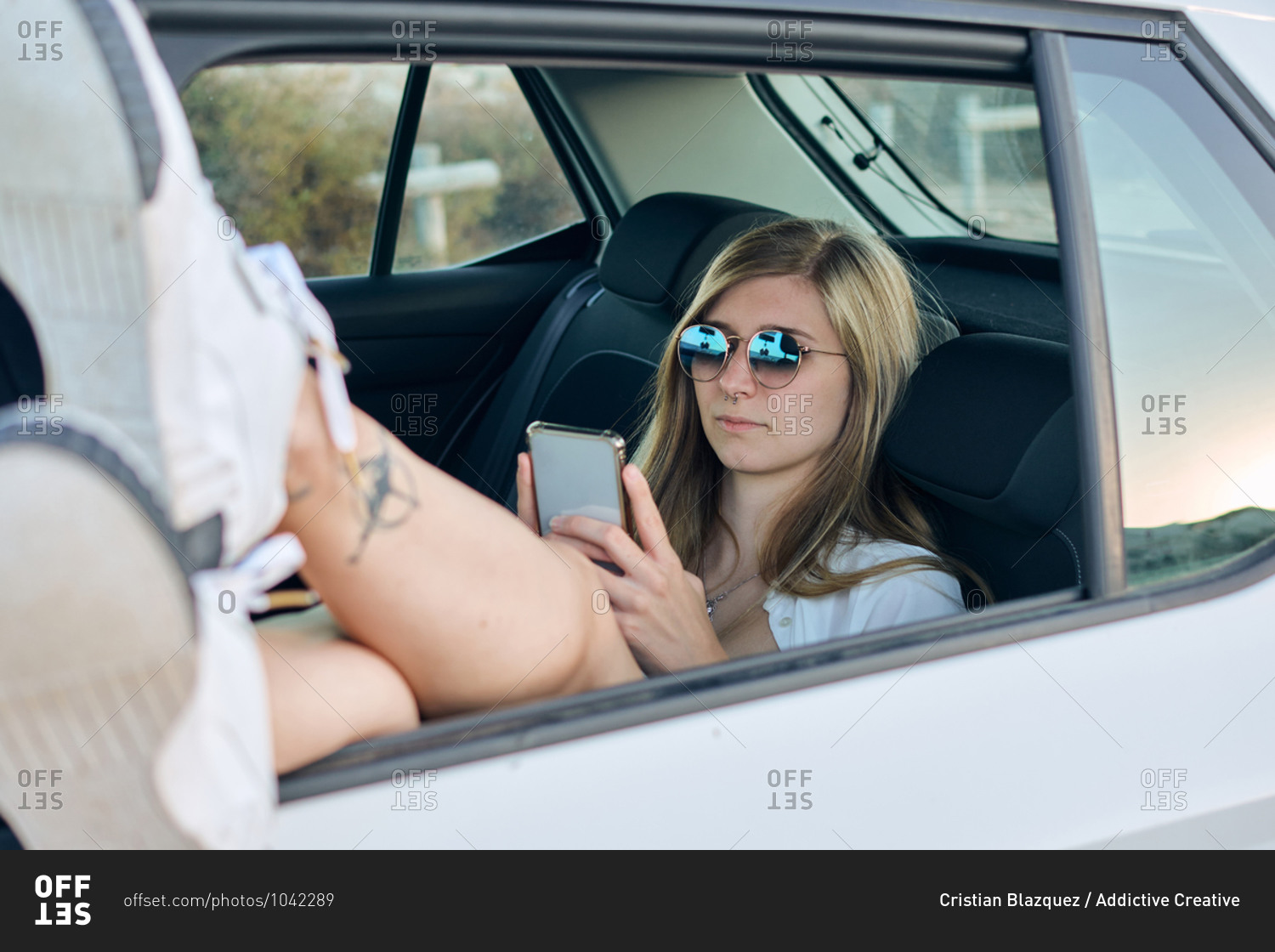 Carefree female traveler resting on backseat of car with legs out of window and reading messages on cellphone during vacation