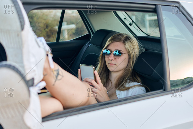 Carefree female traveler resting on backseat of car with legs out of window and reading messages on cellphone during vacation