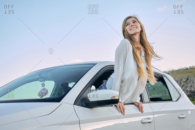 Carefree female driver looking out of window of automobile and enjoying fresh air in nature during summer vacation in evening while looking away