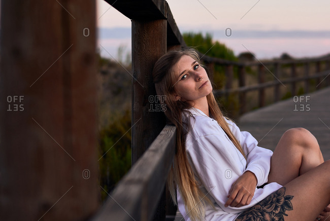 Side view of dreamy female traveler sitting on embankment and leaning on wooden fence while enjoying sunset looking at camera in summer
