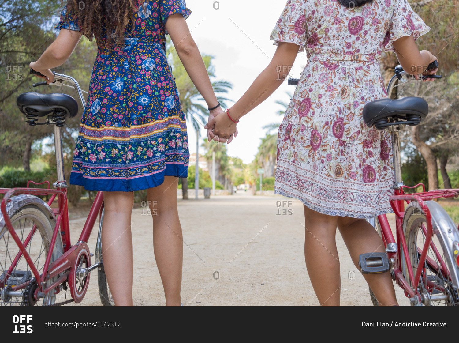 Back view of crop anonymous young female friends in colorful dresses holding hands while standing with bikes on pathway in green tropical park during summer holidays