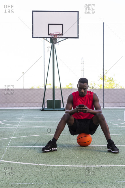 Calm African American sportsman sitting on basketball on playground and texting on mobile phone while looking away