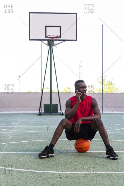 Calm African American sportsman sitting on basketball on playground and speaking on mobile phone while looking away