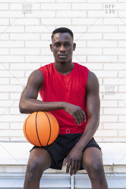Confident African American male basketball player in sportswear sitting with ball on stone border on playground and looking at camera