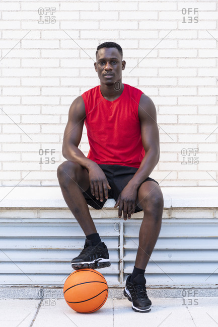 Confident African American male basketball player in sportswear sitting with ball on stone border on playground and looking at camera