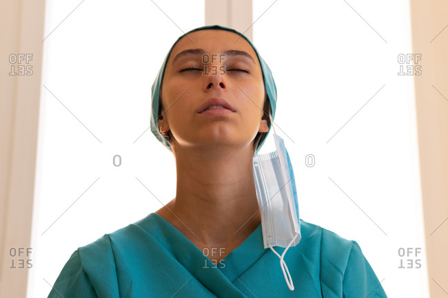 Low angle of tranquil female medic in uniform standing in clinic without mask and breathing in fresh air with closed eyes after coronavirus outbreak