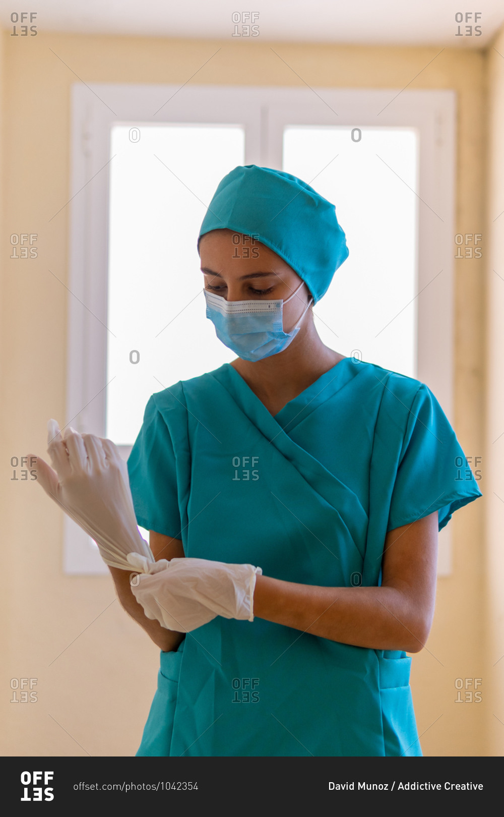 Young nurse in blue scrubs and medical mask putting on sterile latex gloves while preparing for work in clinic during coronavirus pandemic