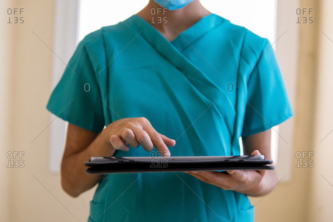 Young nurse in blue scrubs and medical mask reading information on tablet while standing in corridor of modern hospital