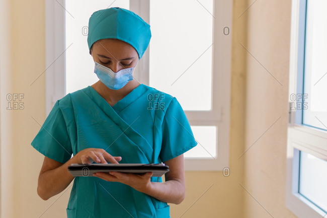 Young nurse in blue scrubs and medical mask reading information on tablet while standing in corridor of modern hospital