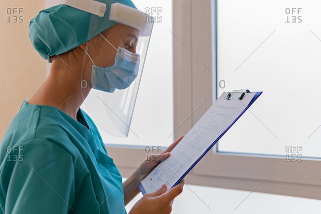 Serious female medic in protective face shield and mask reading clipboard while working in hospital during coronavirus pandemic