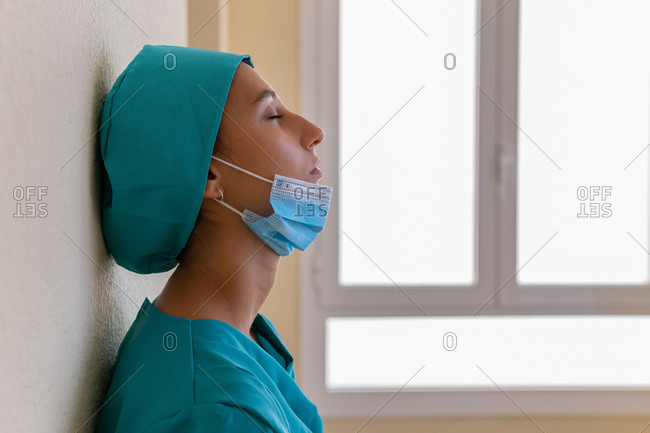 Side view of tired desperate female medic in blue uniform with mask on chin leaning on wall of hospital corridor after working hard during coronavirus pandemic