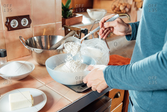 Crop anonymous male adding flour into bowl placed on scales while preparing ingredients for biscuits dough in home kitchen