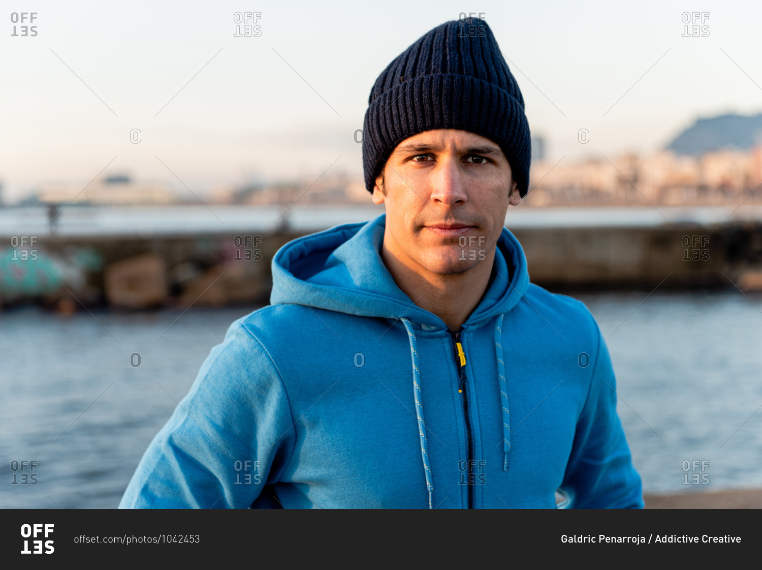 Confident adult ethnic male in warm sportive hoodie and knitted hat looking at camera while standing on embankment near sea at sunset time