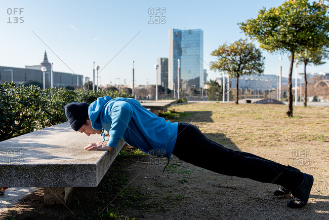 Full body side view of adult sportsman in warm activewear doing push ups exercise against concrete bench during outdoor fitness training in city
