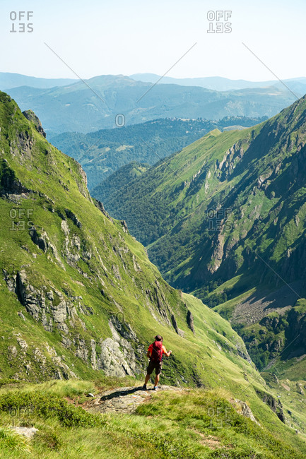 Back view of distant unrecognizable male hiker with backpack and trekking stick standing on grassy hilltop and admiring spectacular scenery of mountain ridge in summer day