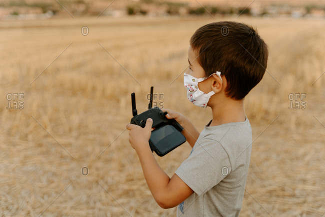 Side view of focused little Asian boy in protective mask using remote control while playing with flying drone in field