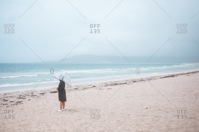 Side view of unrecognizable traveler in raincoat with transparent umbrella walking on empty sandy beach towards waving sea in cloudy day