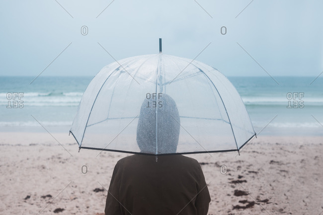 Back view of unrecognizable traveler in raincoat with transparent umbrella walking on empty sandy beach towards waving sea in cloudy day