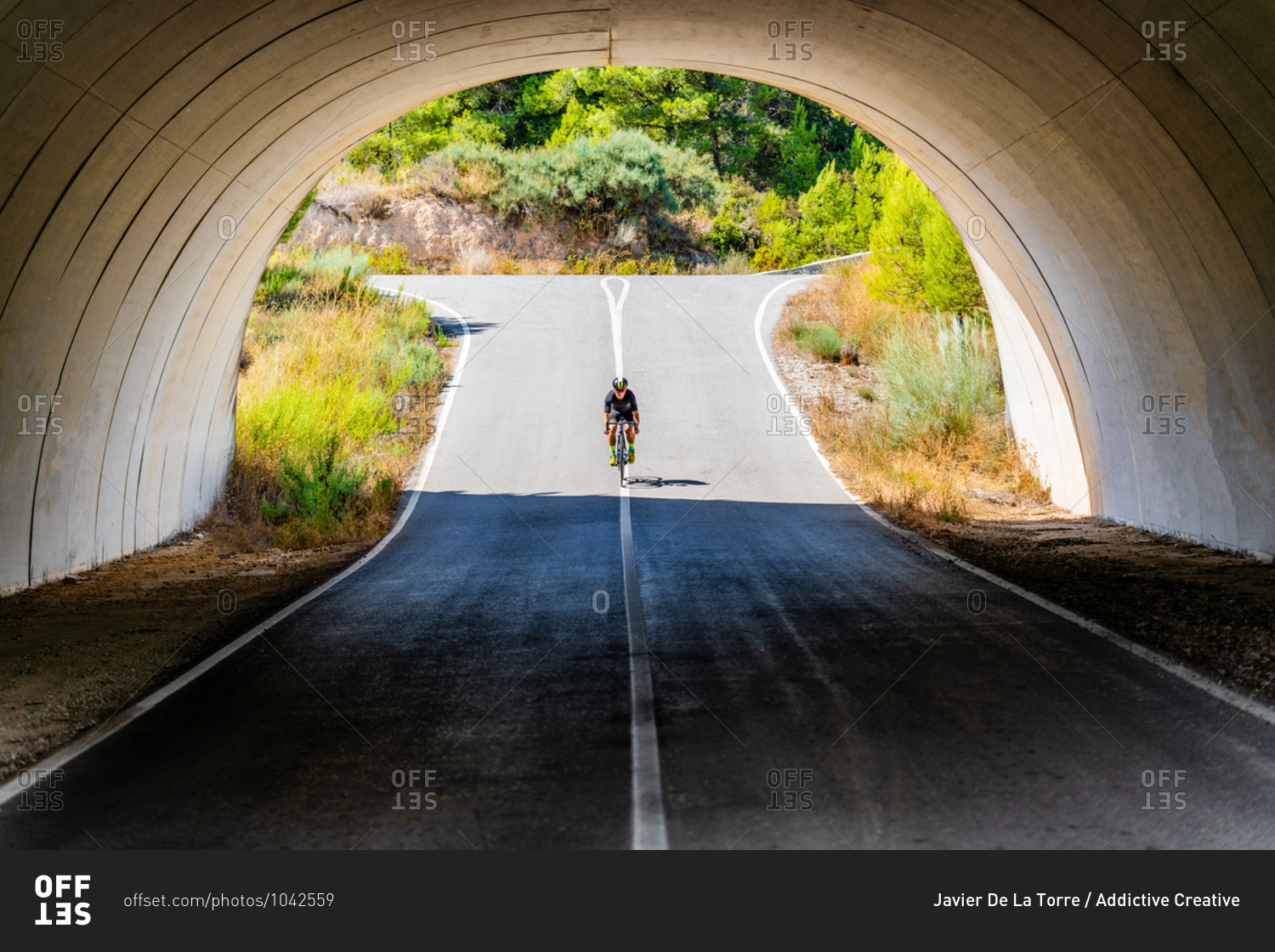 Full length of bicyclist in sportswear and helmet cycling on asphalt roadway under arched bridge in summer countryside
