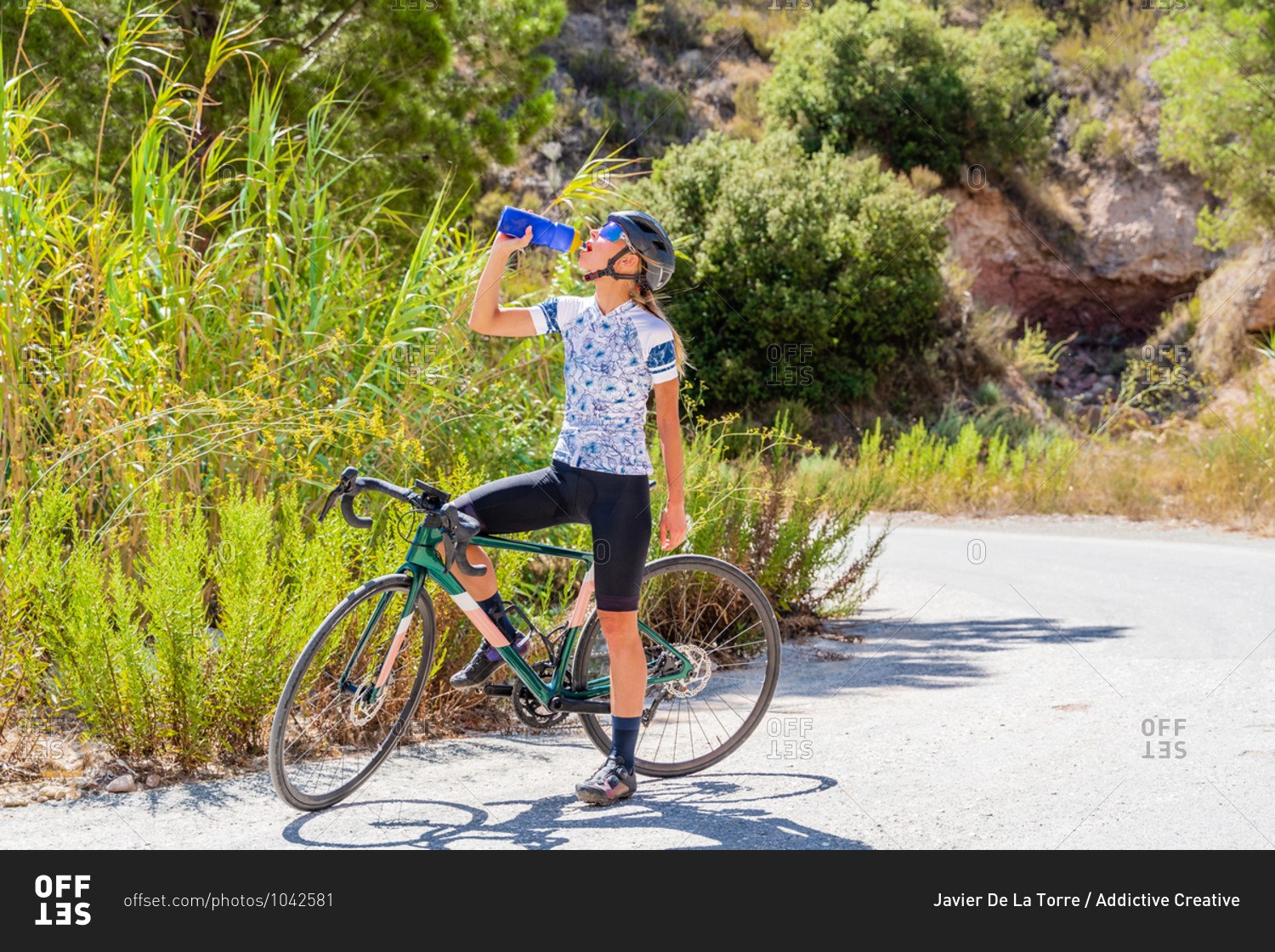 Full body of fit female bicyclist in sportswear and helmet drinking fresh water from bottle while resting after riding bike up mountain road