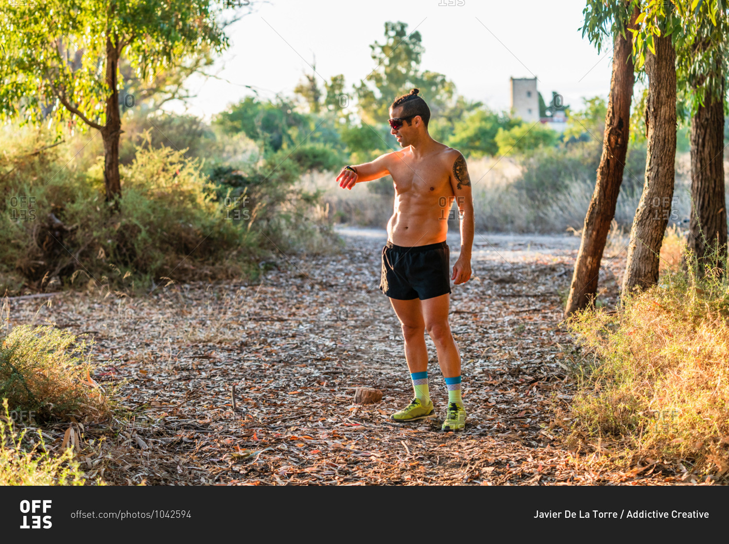 Full body of athletic shirtless male runner standing on pathway in park and checking pulse on fitness tracker during outdoor workout in summer day