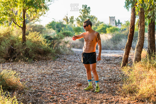 Full body of athletic shirtless male runner standing on pathway in park and checking pulse on fitness tracker during outdoor workout in summer day