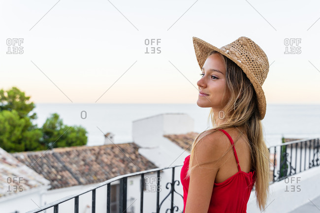 Side view of carefree female tourist standing on terrace and leaning on railing while enjoying amazing cityscape at sunset during vacation in summer