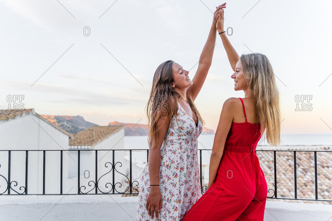 Side view of tender female friends standing on balcony at sunset and holding raised hands while looking at each other and enjoying holiday in summer