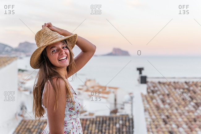 Side view of cheerful female tourist in summer dress and straw hat standing on background of old city near sea and looking at camera at sunset