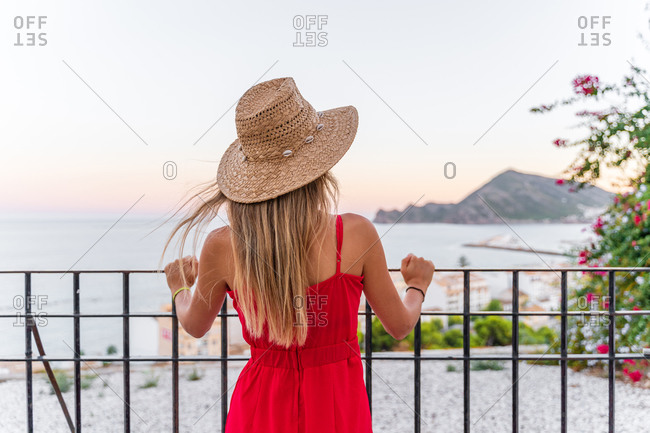 Back view of unrecognizable female tourist standing with straw hat in raised hand on terrace and enjoying summer vacation while admiring amazing cityscape
