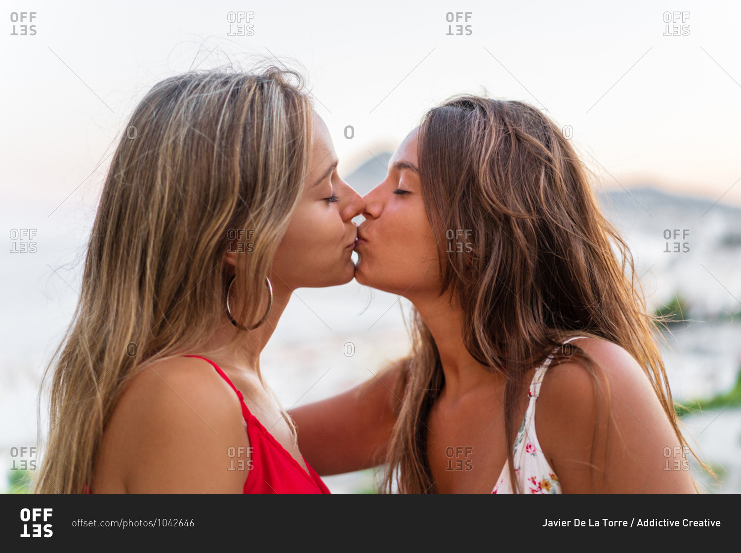 Side view of young girlfriends kissing gently while hugging on background of sundown in summer
